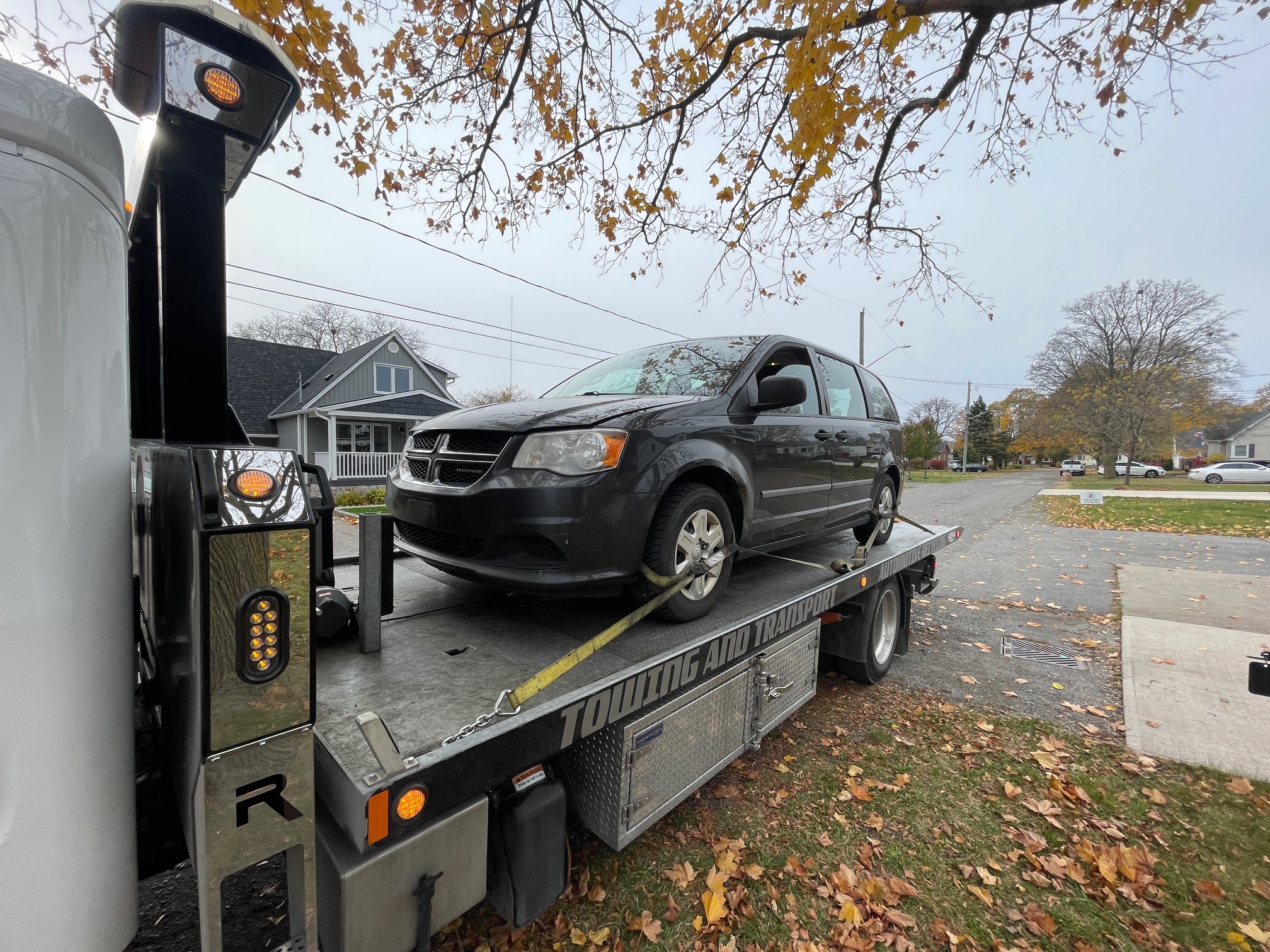 Dodge minivan secured on a flatbed truck on a tree-lined GTA street