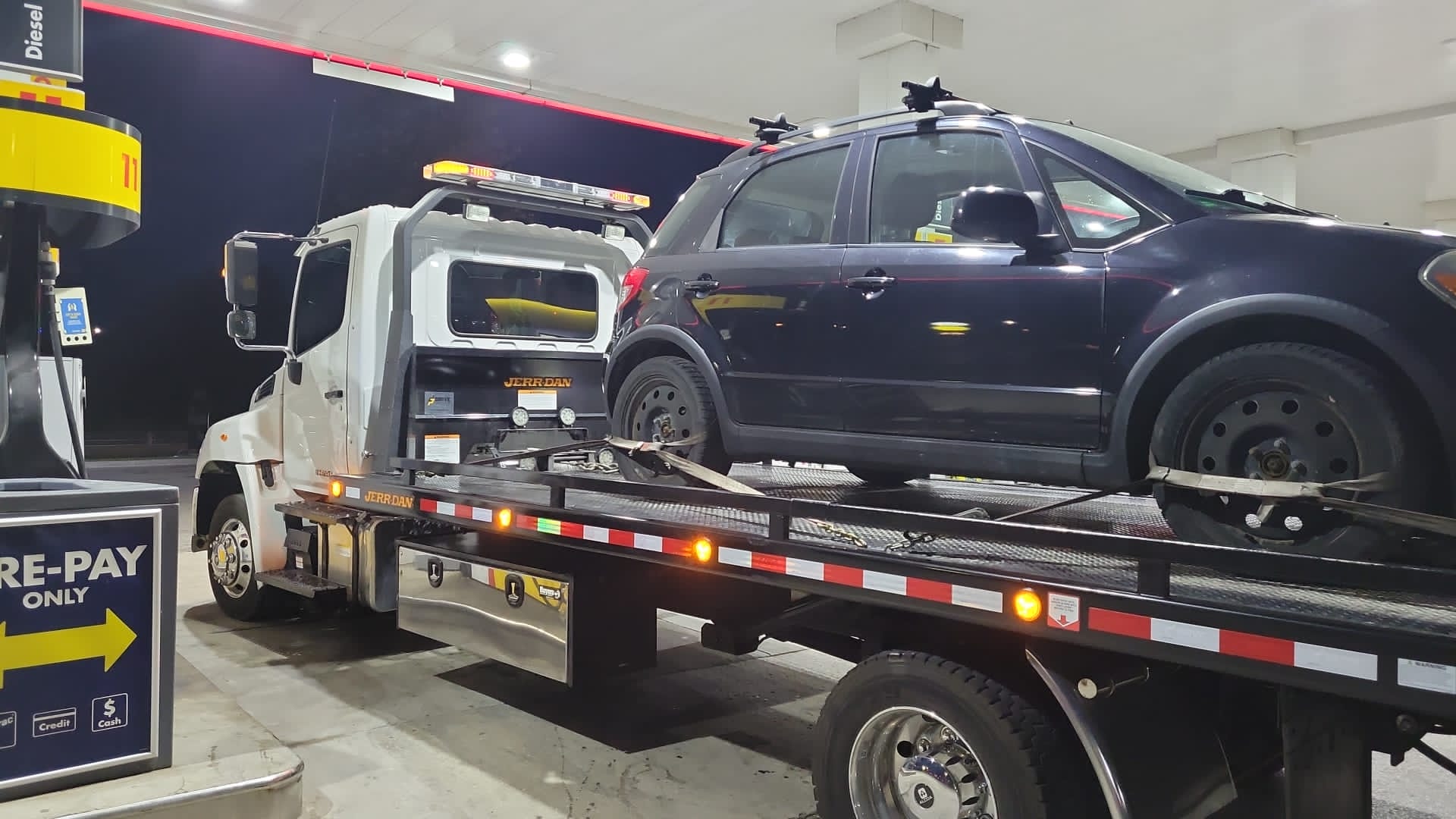 Flatbed tow truck transporting a dark SUV at a Toronto gas station at night